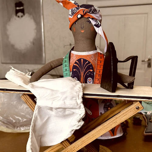A handmade seamstress doll pressing a garment at a miniature ironing board with a vintage flat iron.